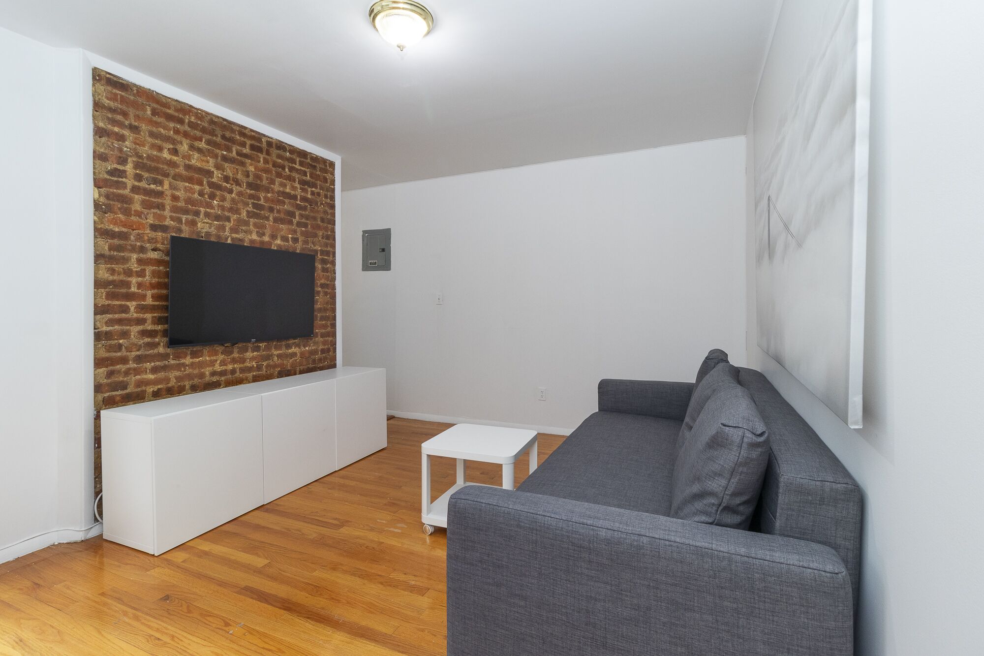 A modern living room with a brick wall, a flat-screen TV, a grey sofa, and a small white table.