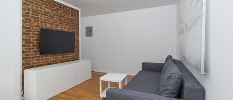 A modern living room with a brick wall, a flat-screen TV, a grey sofa, and a small white table.