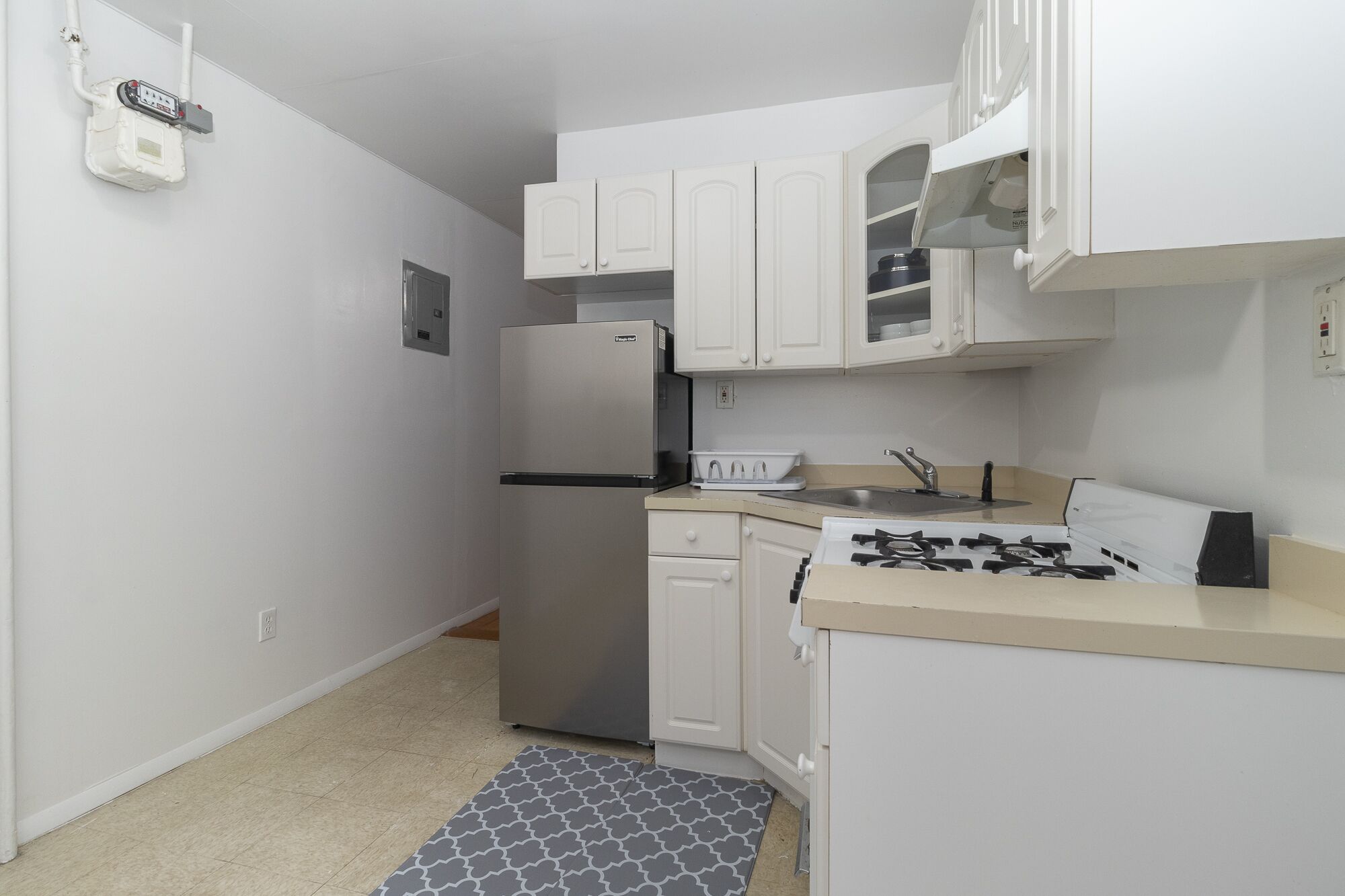 Une cuisine de taille réduite, équipée d’un réfrigérateur, d’une cuisinière, d’un évier et de meubles de rangement blancs.