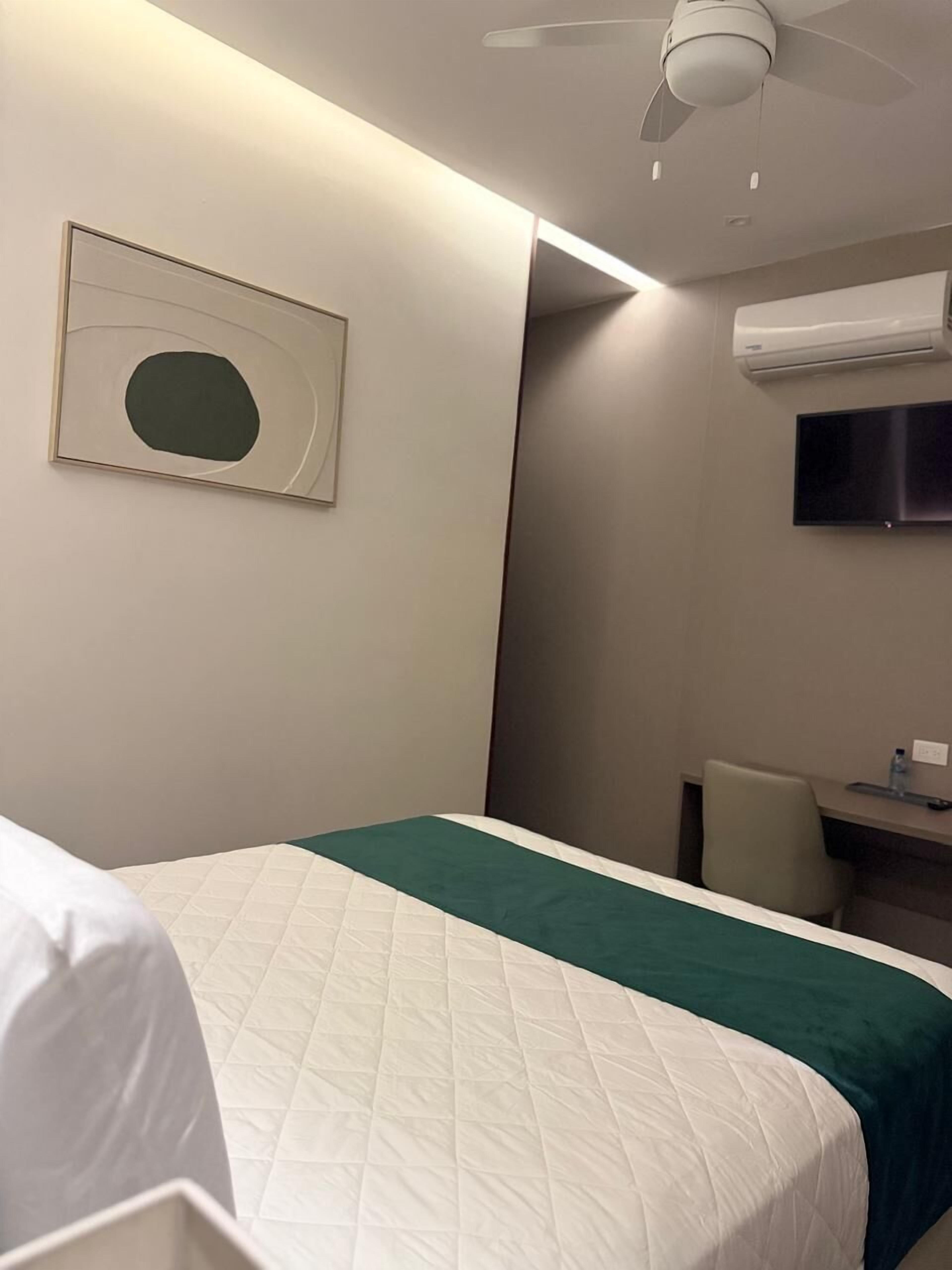 A hotel room with a bed, a desk, a chair, a ceiling fan, an air conditioning unit, and a framed picture on the wall.