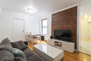 Ein modernes Wohnzimmer mit Backsteinwand, Flachbildfernseher, weißes Sofa und Essbereich mit Tisch und Stühlen.