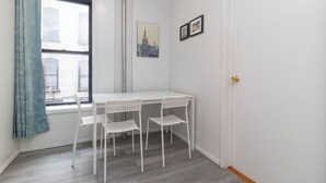 Ein kleiner Raum mit einem weißen Tisch und vier weißen Stühlen, einem Fenster mit Vorhang und zwei gerahmten Bildern an der Wand.