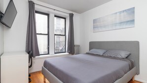 Ein Schlafzimmer mit einem Bett, einem an der Wand montierten Fernseher und einem Fenster mit Vorhängen.