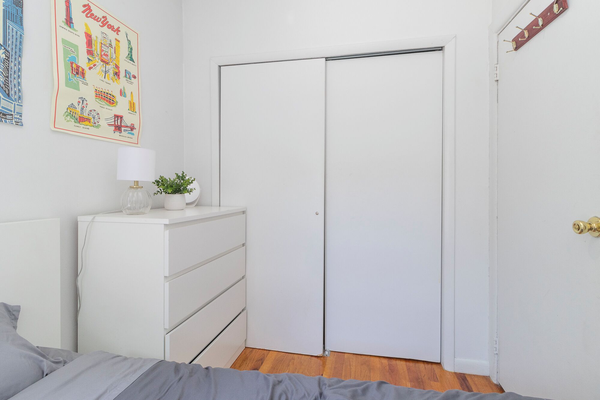 Un dormitorio con un armario blanco, un tocador con una lámpara y una planta, y un cartel llamativo en la pared.