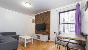 A compact living space with a grey sofa, a white TV stand, a dining table with chairs, and a brick wall.