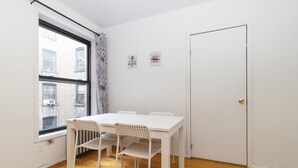 Una habitació petita i lluminosa amb una taula i cadires blanques, una finestra amb vista a edificis, i una porta.