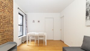 Habitació petita i lluminosa amb una taula i caires blanques, un sofà gris i una paret de maó.
