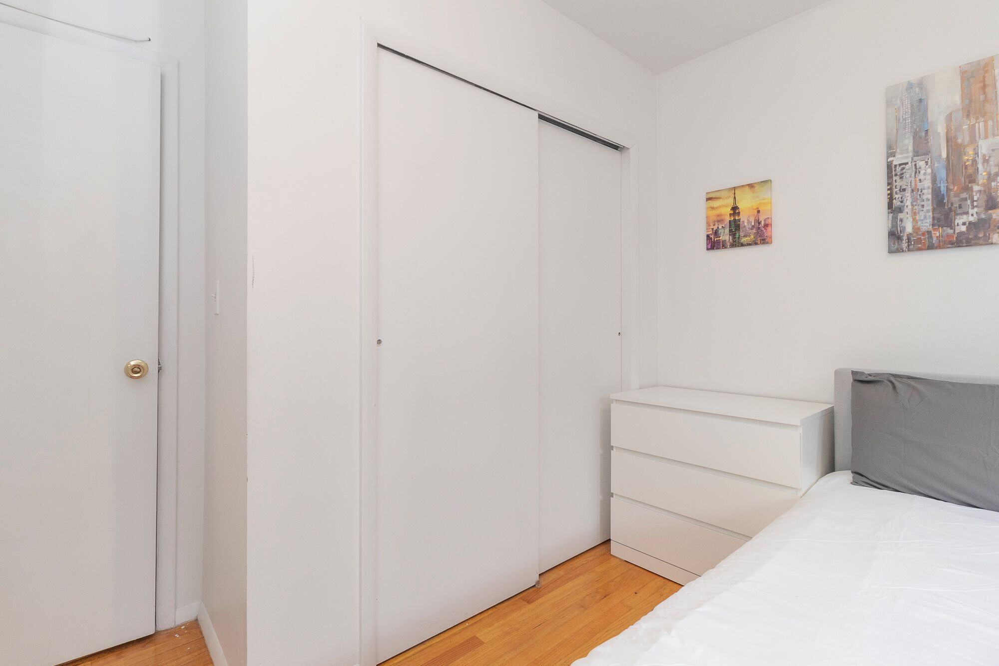 Un dormitorio con un armario blanco, una cama con cabecero gris y dos cuadros en la pared.