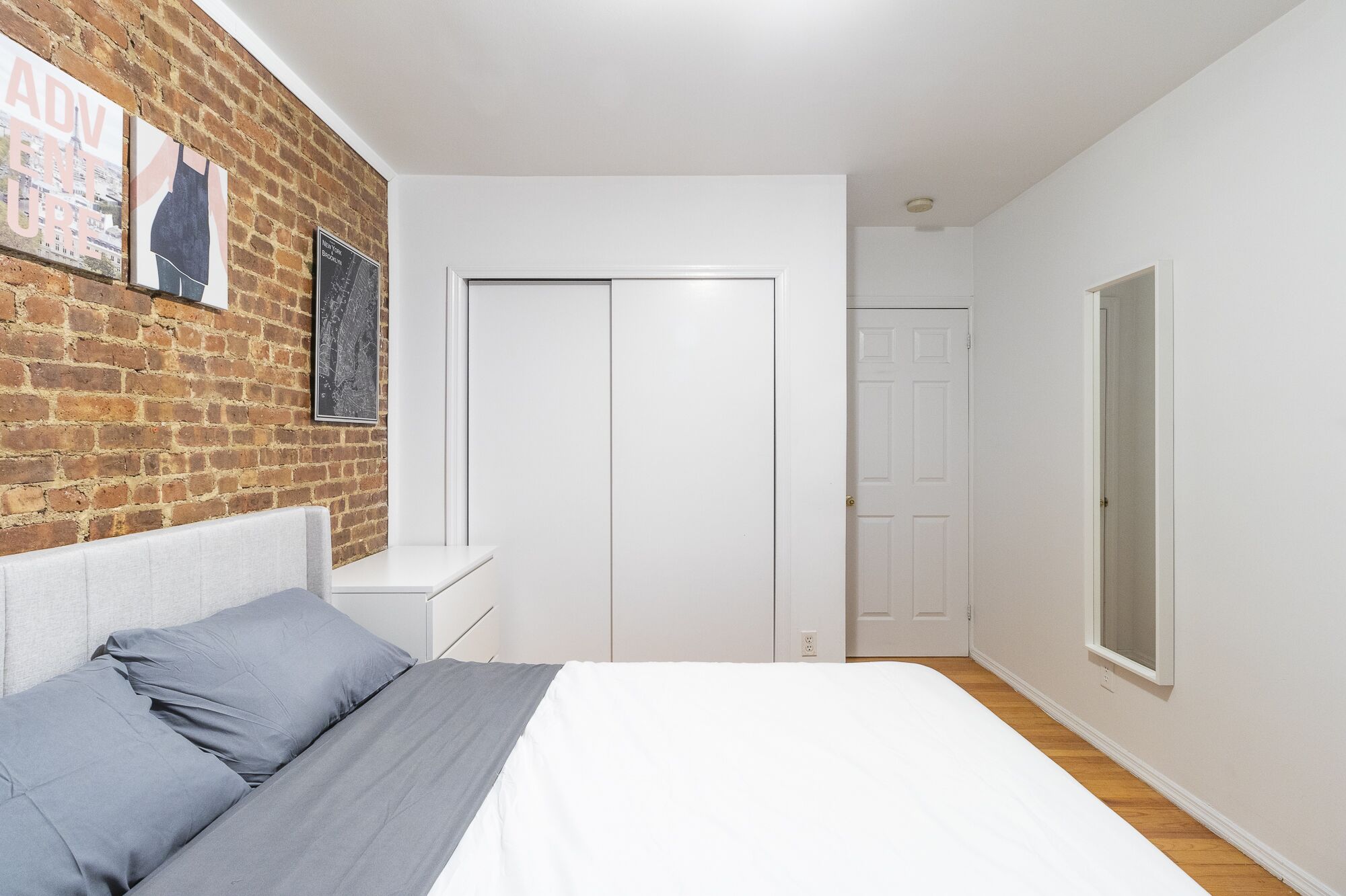 A bedroom with a brick wall, a bed with grey bedding, a white wardrobe, and a window.