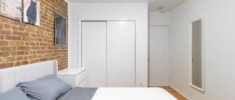 A bedroom with a brick wall, a bed with grey bedding, a white wardrobe, and a window.
