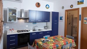 A compact kitchen with blue cabinets, a gas stove, and a sink. There is a dining table with a colorful tablecloth. - Casa Sally with sea view in Macari (San Vito Lo Capo)