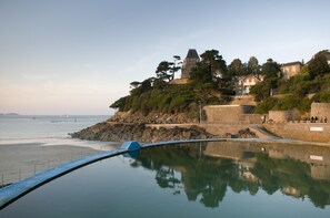 A coastal view with a stone wall, a small tower, and a building with a balcony overlooking the sea.