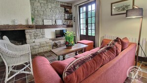 Un salon chaleureux avec une cheminée en pierre, un canapé confortable, un fauteuil en osier et une table à manger agrémentée d’un vase de fleurs.