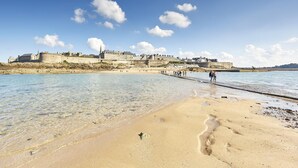 Une plage côtière aux eaux cristallines, avec une jetée et des bâtiments historiques en arrière-plan.
