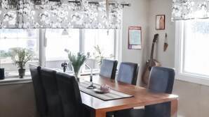Un comedor con una mesa de madera, cuatro sillas y un florero con flores. Hay ventanas con cortinas florales, una guitarra y un cuadro en la pared.