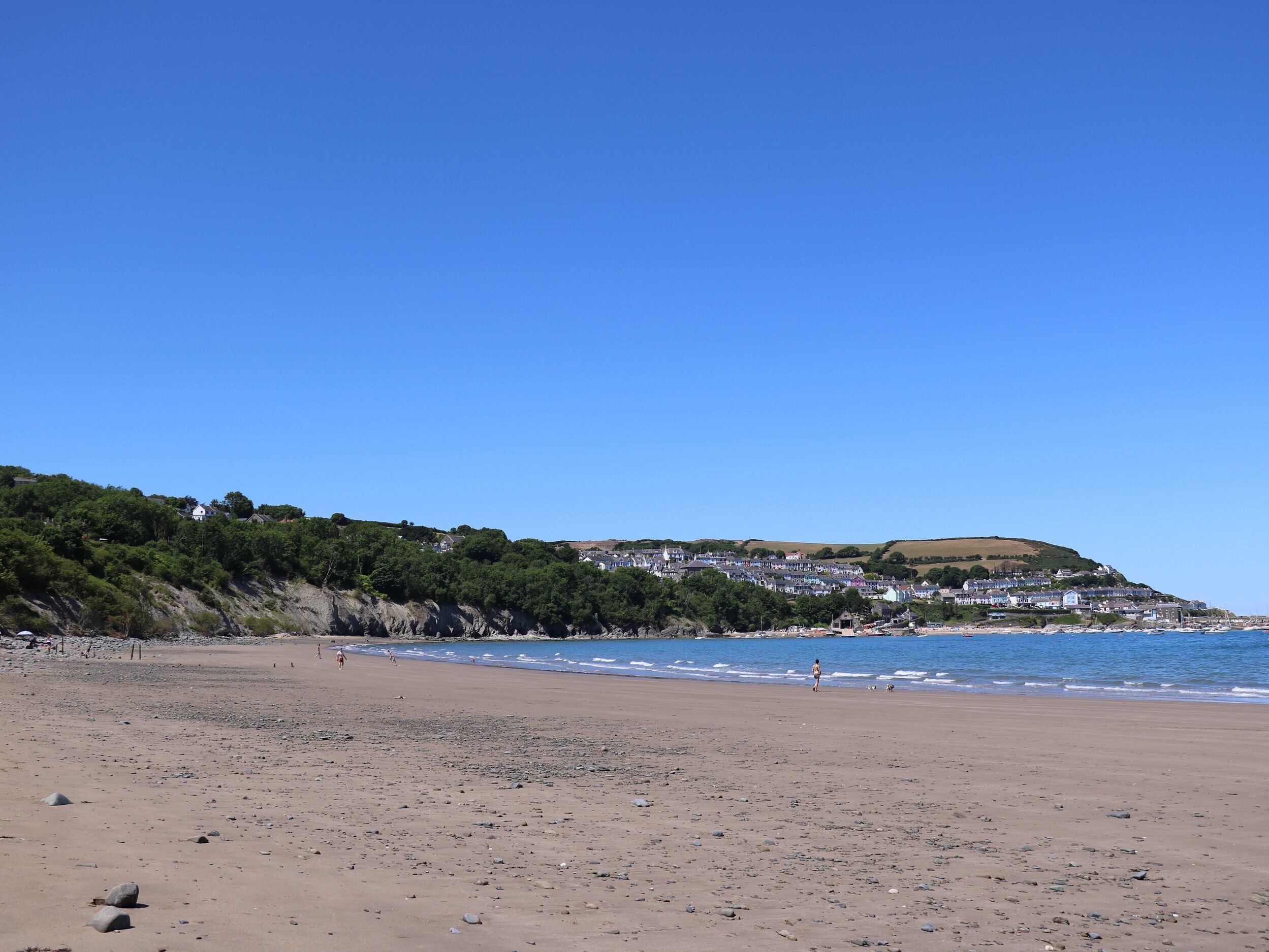 Una playa con costa rocosa, cielo azul despejado y un pueblo en la colina al fondo.