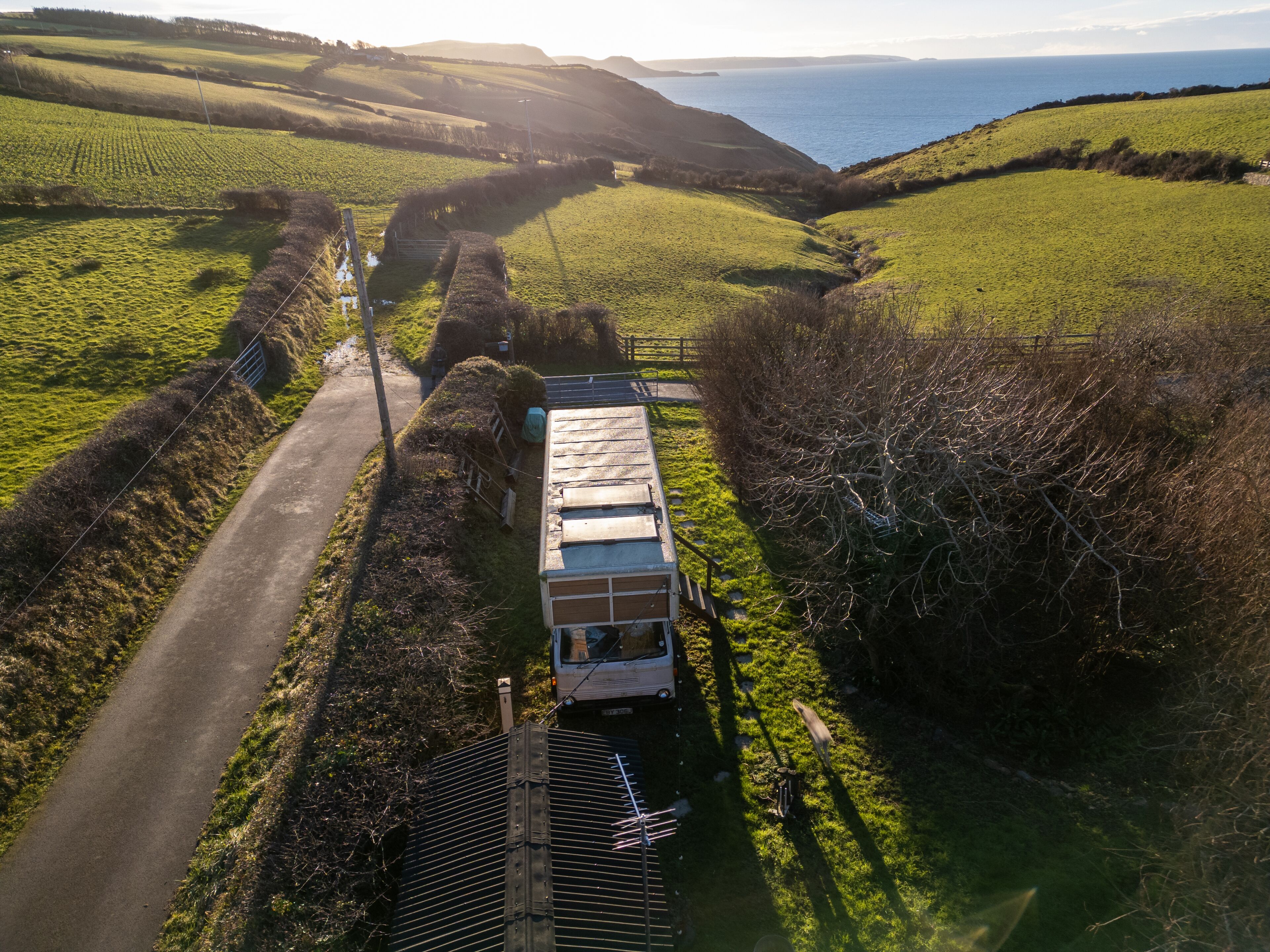 Un paysage rural avec une route, un véhicule blanc et un bâtiment doté d’un toit en pente.