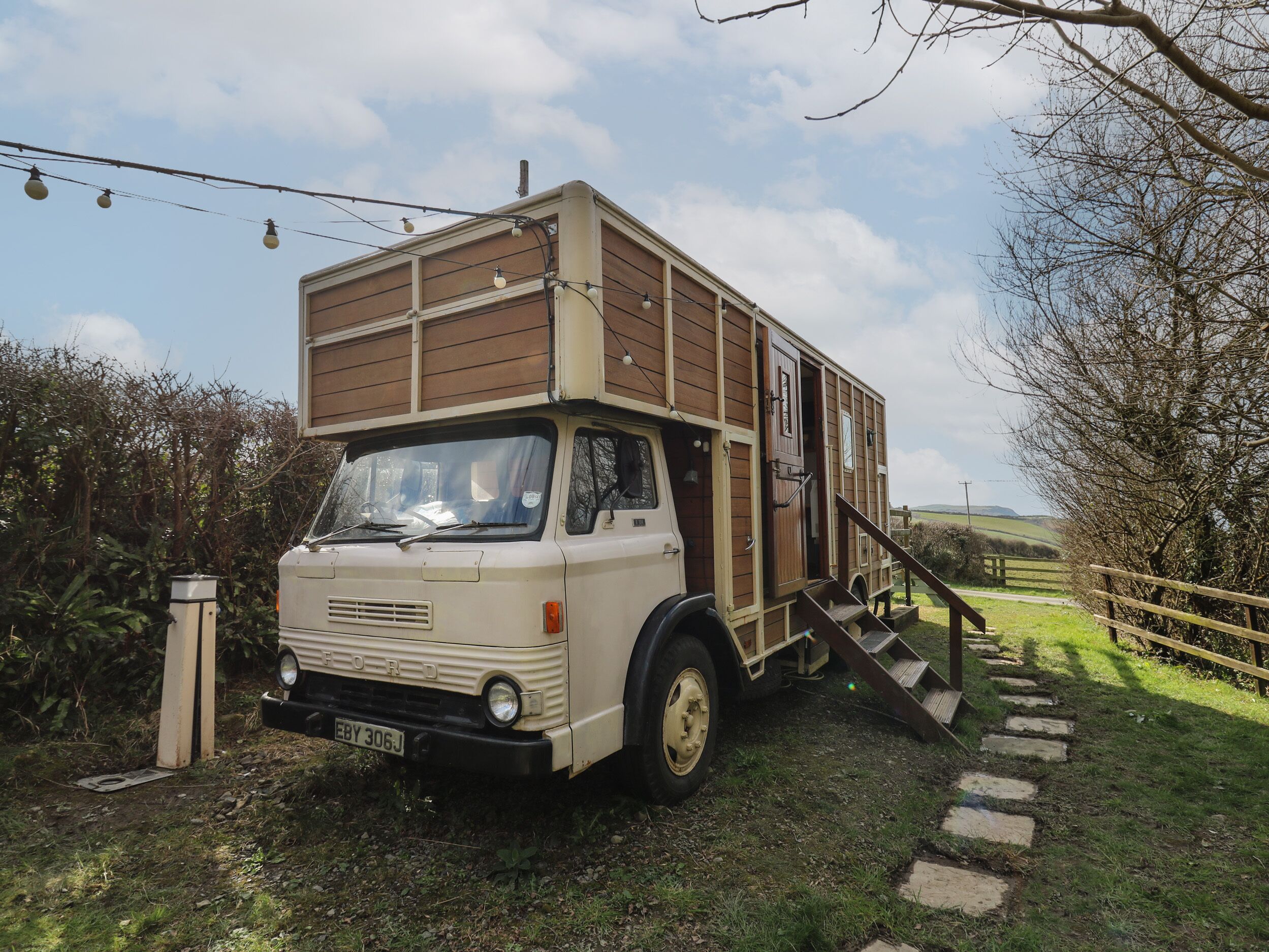 A vintage Ford truck converted into a mobile home with a wooden deck and a staircase leading to the entrance.