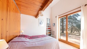 Een slaapkamer met een houten plafond, een bed met een roze en witte dekbedovertrek, een nachtkastje met een lamp, een raam met uitzicht op bomen, en een schuifdeur met glas.