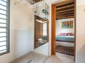 A modern bathroom with a glass shower enclosure, a wooden ceiling, and a bed with a colorful painting in the adjacent room. - Villa Ekinos is ideally located within walking distance of the beaches. (Le Moule)