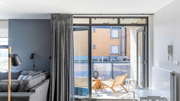 A modern living room with a grey sofa, a large window with curtains, a small table, and a chair.