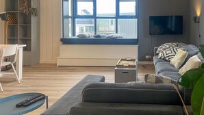 A modern living room with a large window seat, a grey sofa, a flat-screen TV, and a bookshelf.