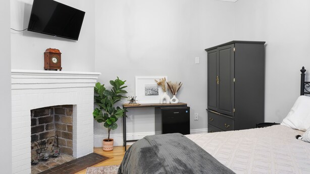 Quarto com lareira, televisão na parede, secretária com computador, cama com cobertor cinza e guarda-roupa escuro.