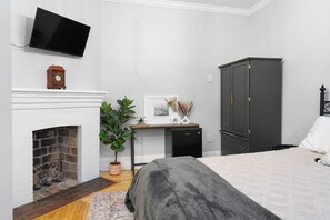 Une chambre avec une cheminée, un téléviseur fixé au mur, un bureau avec un ordinateur, un lit avec une couverture grise et une armoire de couleur sombre.