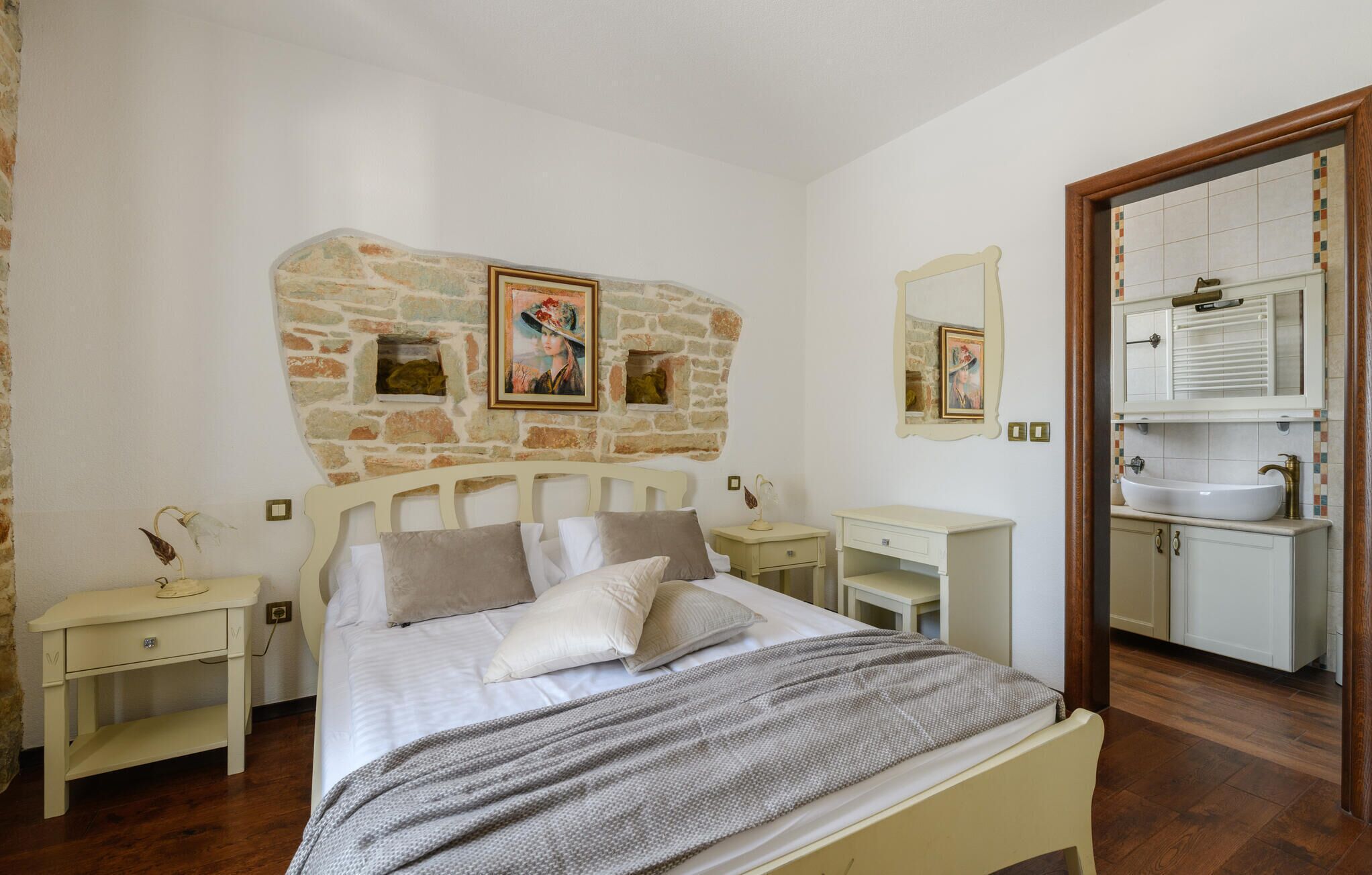 A bedroom with a stone wall, a bed with pillows, a bedside table, a nightstand, a mirror, and a bathroom with a sink and cabinet.