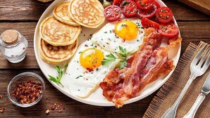 A breakfast plate with eggs, bacon, tomatoes, olives, and pancakes on a wooden table.