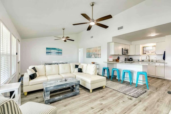 A modern living room with a sectional sofa, a coffee table, and a kitchen area with bar stools. - Surfside Beach House: Steps to the Sand & Sun! (Surfside Beach)