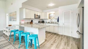 Una cocina moderna con armarios blancos, encimera de granito y refrigerador de acero inoxidable. Hay tres taburetes azules y una ventana con vista a la playa.