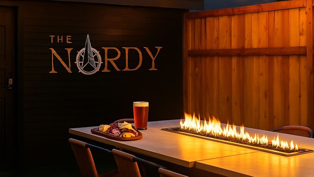 A wooden table with a fire feature, a glass of beer, and a plate of food. The wall has a logo that reads "THE NORDY".