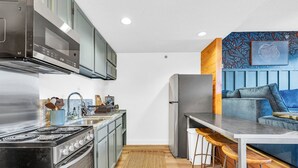 Cozinha compacta com fogão, forno, micro-ondas e geladeira. Há um espaço de bar com banquetas e um sofá azul.