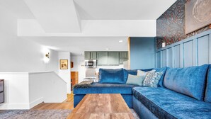 Uma sala de estar moderna com um sofá modular azul, piso de madeira e uma cozinha ao fundo.