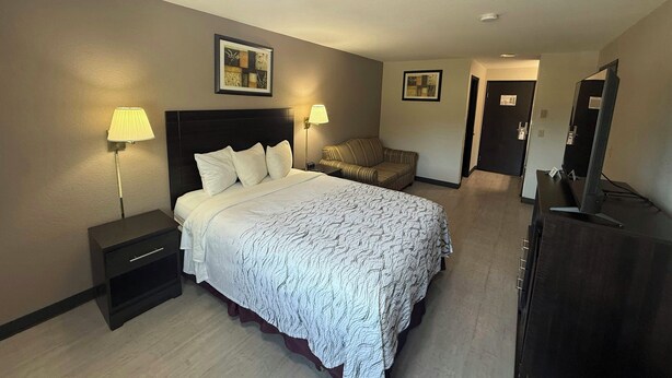 A hotel room with a bed, bedside tables, a lamp, a chair, a dresser, and a mirror. - Red Roof PLUS+ Centralia (Centralia)