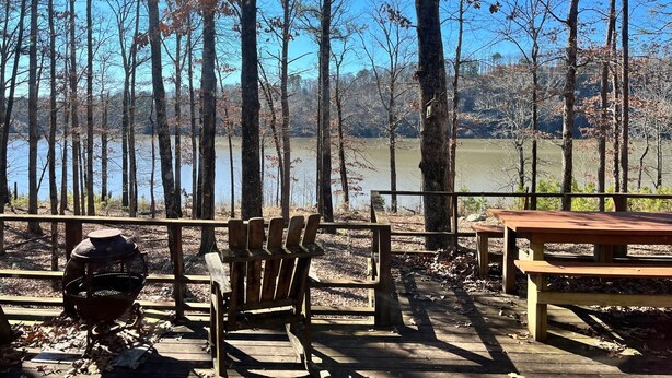 Una terraza de madera con una parrilla, sillas y una mesa de picnic, con vistas a un lago rodeado de árboles.