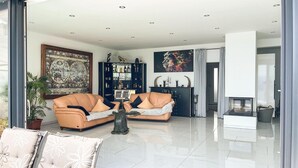A modern living room with a sofa set, a coffee table, and a fireplace. There is a large painting on the wall and a bookshelf filled with books and decorative items.