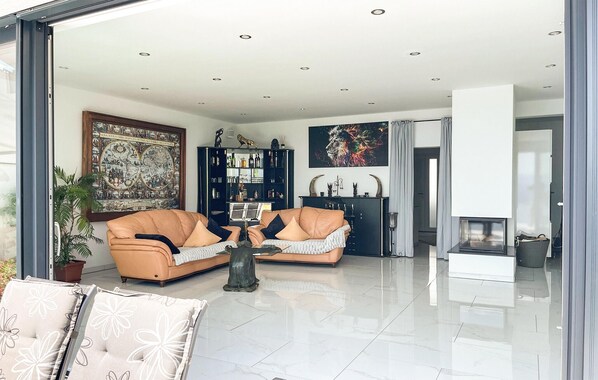 A modern living room with a sofa set, a coffee table, and a fireplace. There is a large painting on the wall and a bookshelf filled with books and decorative items.