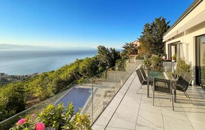 Terraço com guarda-corpo de vidro oferecendo uma vista panorâmica do mar e colinas.