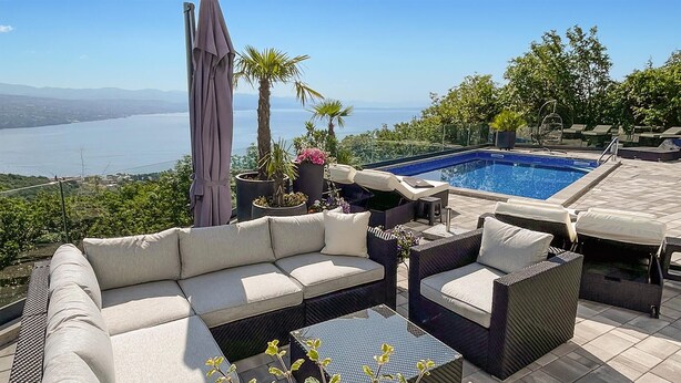 A poolside area with a sofa, chairs, and a table, overlooking a body of water and hills.