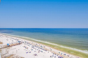 A beach with numerous umbrellas and sun loungers, a pier extending into the water, and a clear blue sky. - A Wave From It All - 19th Floor Amazing Views (Gulf Shores)