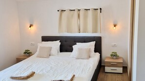 Un lit bien fait, avec du linge de lit blanc, deux tables de chevet ornées de petites plantes en pot et une tête de lit rembourrée de couleur sombre.