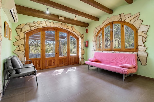 Ein Zimmer mit steinernen Rundbogenfenstern, einem rosafarbenen Sofa, einem grauen Sofa und einem Deckenventilator.
