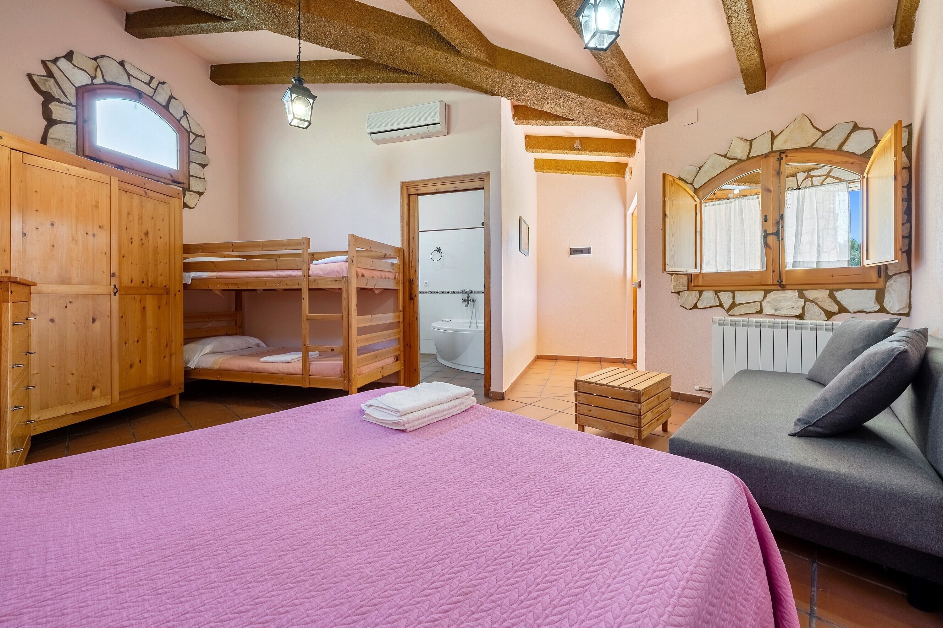Une chambre avec un lit superposé, un lit simple recouvert d’une couette rose, une armoire en bois, un canapé gris, une fenêtre encadrée de pierres et une arche en pierre au-dessus de la porte.