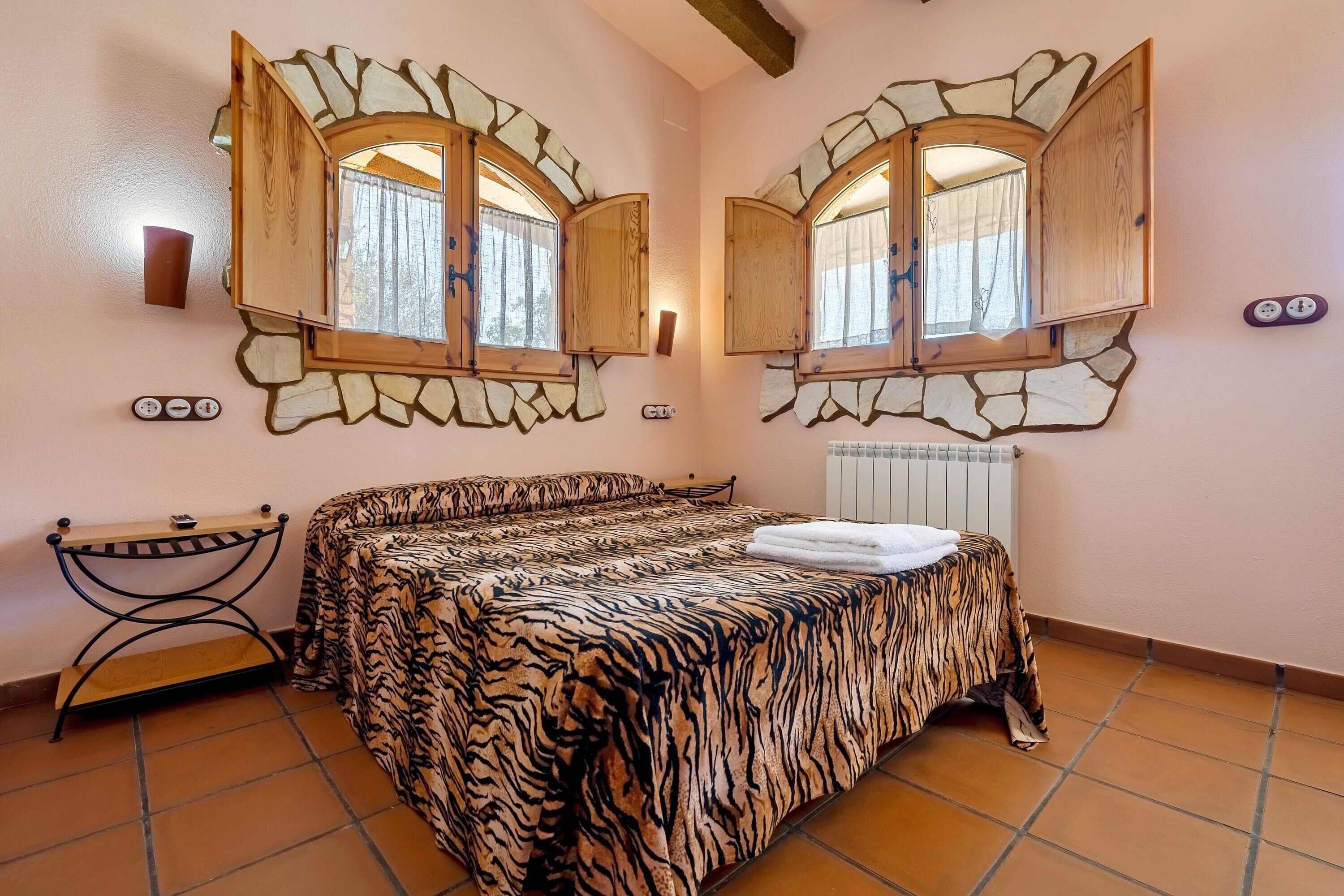 Ein Schlafzimmer mit Bett, zwei Fenstern mit Holzrollläden, einem Heizkörper und einem kleinen Tisch.