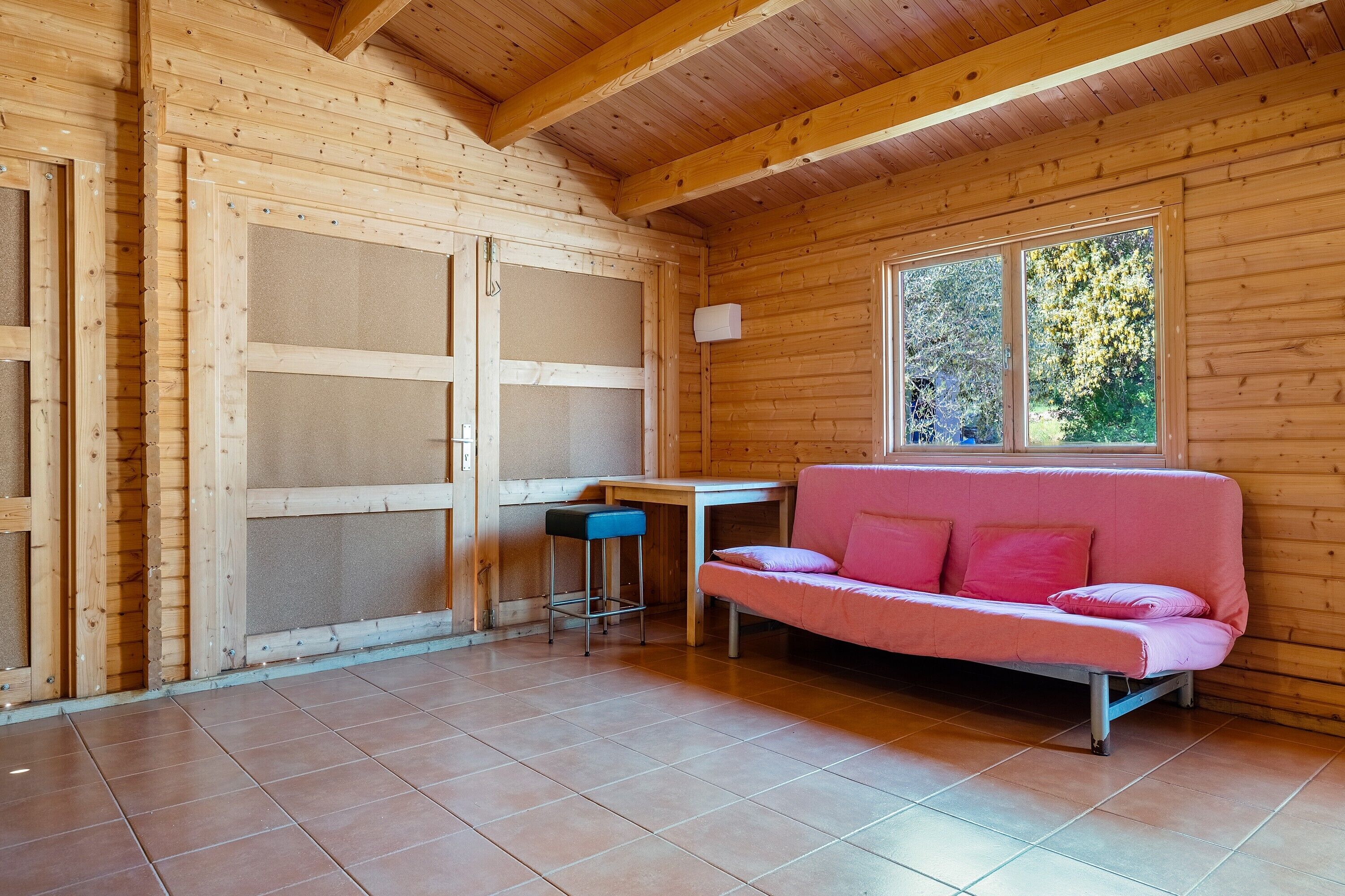 L’intérieur d’un chalet en bois, avec un canapé rose, une petite table et une fenêtre donnant sur les arbres.