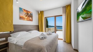 Ein Hotelzimmer mit Bett, einem Sonnenuntergangsbild, Meerblick und einem Balkon mit gelben Vorhängen.