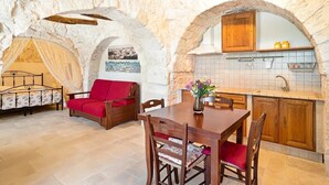 Une pièce aux murs de pierre, comprenant une table à manger, des chaises, un canapé rouge, un lit et un coin cuisine avec des meubles en bois et un pare-feu en carreaux.