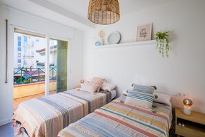 Quarto com duas camas, planta pendente, prateleira na parede com objetos decorativos e vista para a varanda.